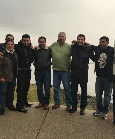 A group of eight men, dressed casually, standing together by a water body under a cloudy sky. The atmosphere appears relaxed and friendly.