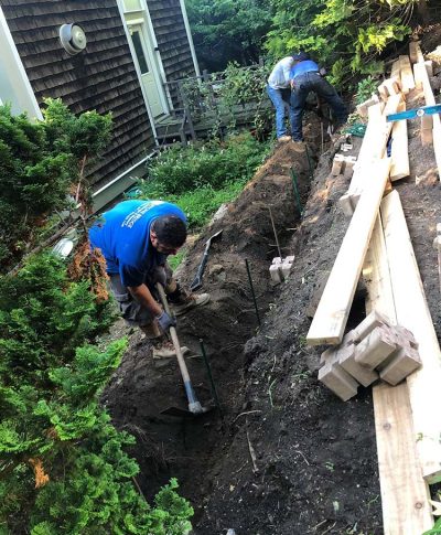 Two workers dig a trench in a garden, surrounded by trees and lumber, to prepare for a construction project.