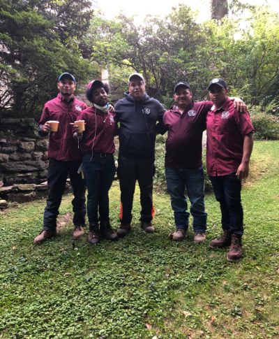 Five people stand together outdoors, wearing maroon shirts and boots, holding drinks, with greenery in the background.