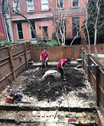 Two workers in red shirts are digging and leveling a garden bed in a fenced backyard, surrounded by leafless trees and brick buildings.